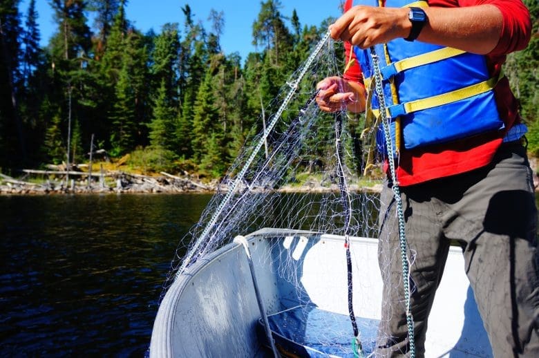 Man in blue life jacket stands on boat in a lake with a net in his hand dangling into the lake.