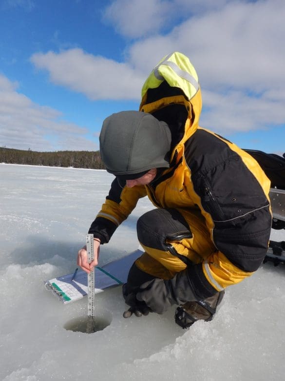 Scientists kneels down on a frozen lake while measuring a hole in the lake using a ruler.