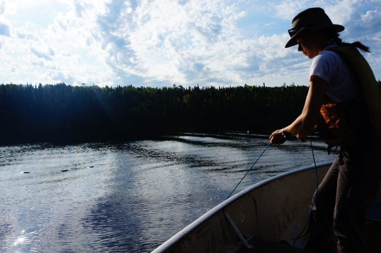 Woman stands in boat and holds a trap net over the edge in a freshwater laker bordered by trees.