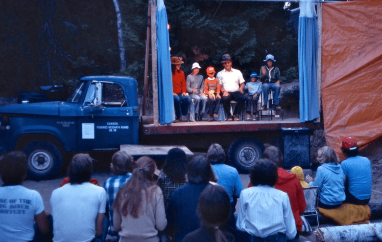 Group of people with back to camera watch people on a stage on a trailer sitting in a straight line