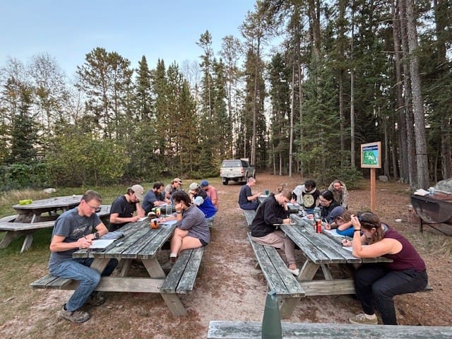 People sitting at lunch desks writing on pads in front of trees