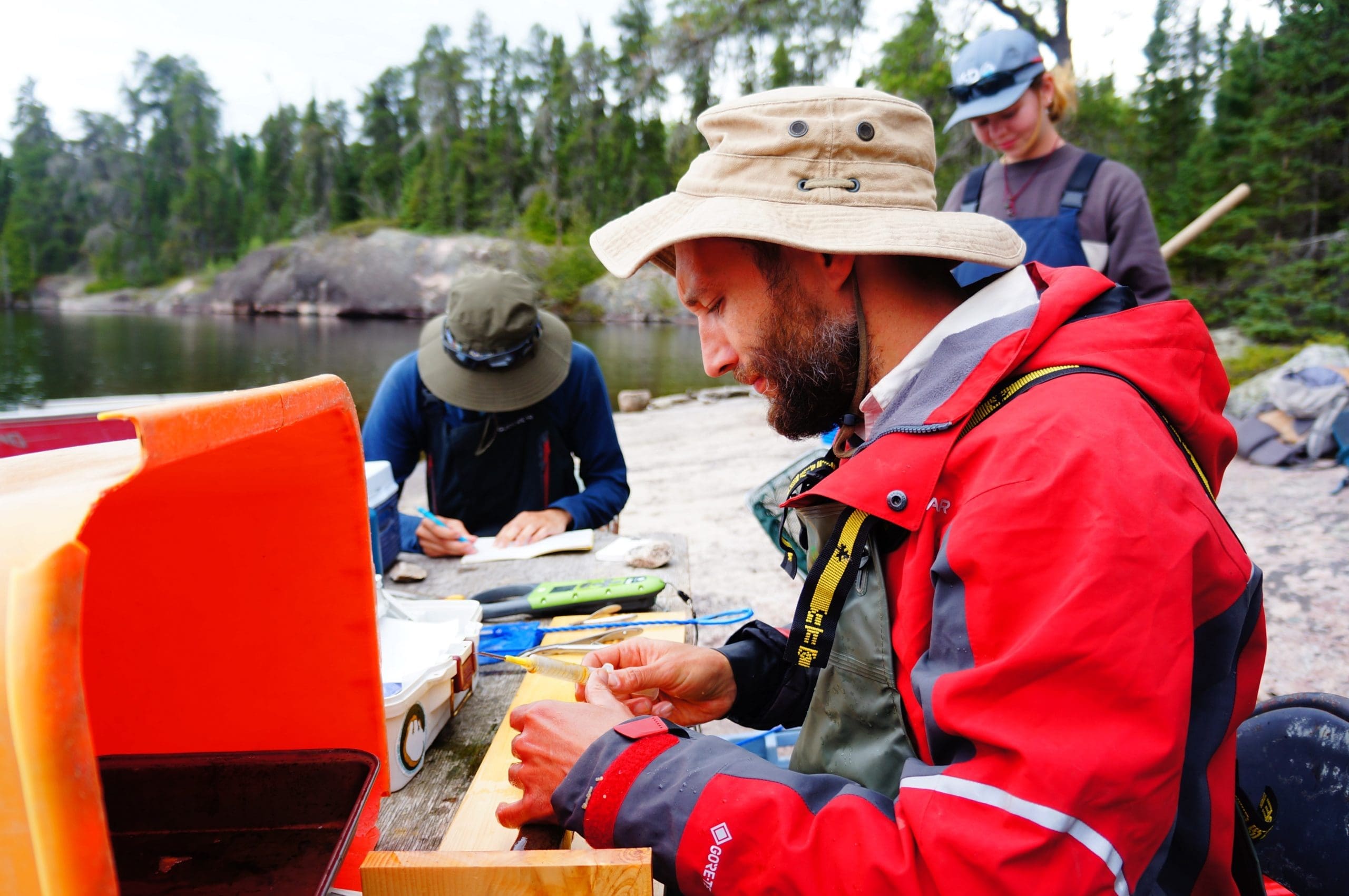 scientist with a red jacket and wide-brimmed hat sits at a table with fish in front of a lake